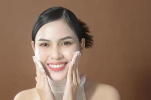 una mujer joven con cara hermosa sonriendo, lavándose la cara sobre fondo marrón, concepto de cuidado de la piel de belleza foto