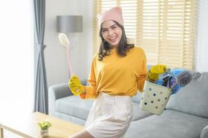 joven mujer feliz usando guantes amarillos y sosteniendo una canasta de artículos de limpieza en la sala de estar. foto