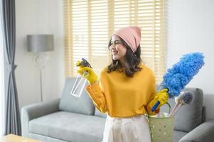 joven mujer feliz usando guantes amarillos y sosteniendo una canasta de artículos de limpieza en la sala de estar. foto