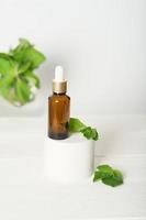 Mock up glass dropper bottle on a white podium on a white background, with mint leaves. Cosmetic serum product on a white background. vertical with mint leaves photo