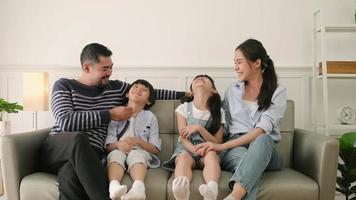 ritratto di famiglia tailandese asiatica, papà adulto, mamma e bambini piccoli che vivono a casa di felicità, relax e presa in giro insieme, tempo libero sul divano nella stanza bianca, fine settimana incantevole e benessere stile di vita domestico. video