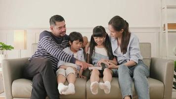 família tailandesa asiática, pai adulto, mãe e filhos felicidade em casa vivendo atividades relaxantes e lendo livro juntos, lazer no sofá na casa do quarto branco, fim de semana adorável, estilo de vida doméstico de bem-estar. video