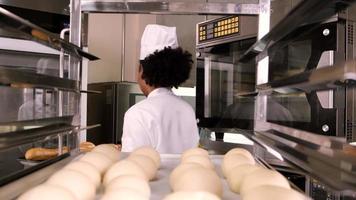 professionele Afro-Amerikaanse vrouwelijke chef-kok in wit kookuniform met hoed en schort die brood maakt van gebakdeeg, vers bakkerijvoedsel bereidt, in de oven bakt in de roestvrijstalen keuken van het restaurant. video