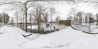 Winter full spherical seamless hdri panorama 360 degrees angle view in snowy park near the bridge of small river with gray pale sky  in equirectangular projection. VR AR content photo