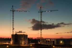 Silhouette tower cranes and unfinished multi-storey high buildings under construction at the sunset in desert on illuminated building site photo