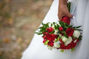 bride in a white dress with a wedding bouquet of white and red roses in park photo