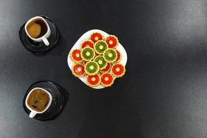 two cups of morning coffee and a plate of colored cookies on a black wooden table. Top view photo