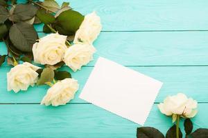 Frame of white roses on blue wooden background with paper card and copy space. Top view and selective focus. Mock up photo