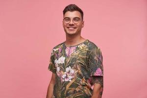 Studio shot of beautiful young tattooed male with charming smile posing over pink background, looking happily to camera, wearing glasses and flowered t-shirt photo