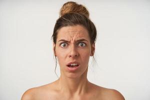 Indoor portrait of dazed young attractive woman with open-eyed face, wrinkling forehead with opened mouth, posing over white background without make-up photo