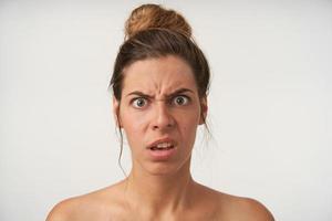 Studio shot of perplexed young female posing over white background with wide eyes opened, frowning and grimacing, wearing bun hairstyle and no make-up photo