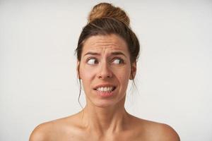 Studio shot of beautiful young woman posing over white background without make-up, looking aside with doubting face, contracting brow and showing teeth photo