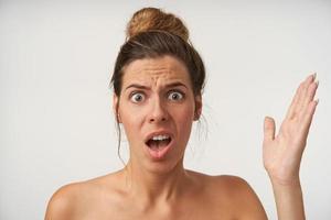 Indoor portrait of confused attractive young woman standing over white background with raised palm, frowning with wide eyes and mouth opened photo