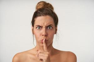 Indoor shot of pretty grumpy female raising index finger to her lips, asking to keep silence, frowning and looking seriously to camera photo