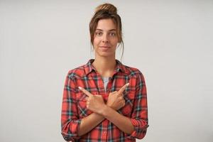 Young attractive woman posing over white background with crossed hands on her chest and pointing with forefingers to different sides, looking to camera with calm face photo