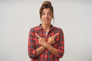 Portrait of young pretty woman in checkered shirt crossing hands on her chest and pointing with forefingers to different sides, biting underlip and contracting forehead photo