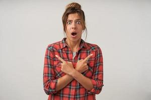 Indoor shot of shocked pretty woman with crossed hands on her chest, looking to camera with wide opened mouth and pointing with index fingers to different sides photo