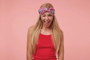 hermosa joven rubia feliz posando sobre fondo rosa, sonriendo y guiñando un ojo a la cámara, usando una diadema de color y una camisa roja foto