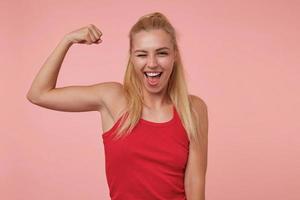 Indoor portrait of sporty young female with casual hairsyle showing strong biceps, looking at camera joyfully and winking, wearing red shirt photo