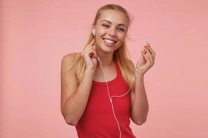 Happy attractive young female with long blonde hair sticking out headphones while listening to music, wearing casual clothes, isolated over pink background photo