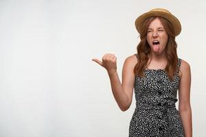 foto de estudio de una joven de pelo rojo con sombrero de paja y vestido informal posando sobre fondo blanco, mostrando con el pulgar a un lado, haciendo muecas y sacando la lengua