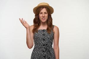 Beautiful young curly woman with red hair looking at camera surprisedly, raising up palm with puzzled face, wearing romantic dress and hat, isolated over white background photo