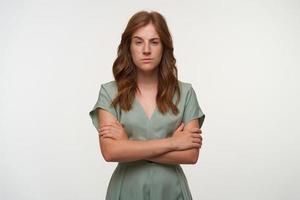 Serious young pretty lady with red hair posing over white background with crossed arms, looking to camera with raised eyebrow, having doubts photo