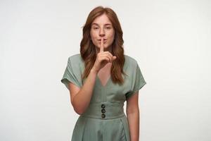 Portrait of beautiful young redhead female in vintage pastel dress posing over white background, raising index finger to lips, asking to keep secret photo