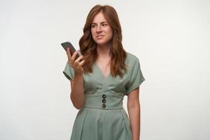 Puzzled pretty female with curly red hair holding smartphone in hand, looking at sreen with confused face, posing over white background photo