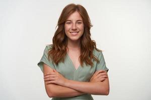 Studio close-up with lovely redhead curly female standing over white backgroung with crossed arms, looking to camera with charming smile photo