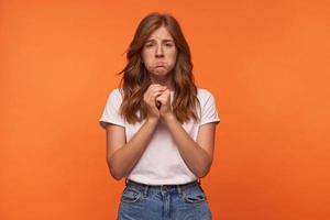 Portrait of unhappy young woman with red curly hair in blue jeans and white t-shirt posing over white background, looking to camera with sad face and raising folded hands photo