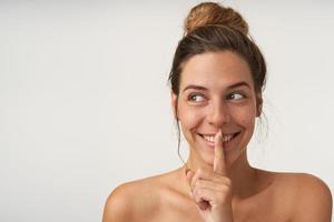 retrato de una joven alegre mirando a un lado con una amplia sonrisa, fingiendo mantener el secreto, manteniendo el dedo índice cerca de los labios, aislada sobre fondo blanco foto