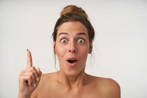 Indoor photo of excited young woman looking at camera with wide eyes opened, raising index finger, having idea and gesturing, isolated over white background