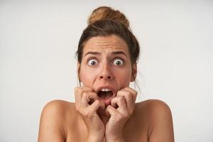 Studio close-up of scared pretty young woman holding hands on face, looking fearful with wide eyes and mouth opened to camera, wearing no make-up photo