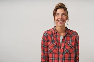 Happy young woman with casual hairstyle posing over white background in checkered shirt, looking aside cheerfully with broad sincere smile photo
