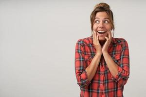 Cheerful young woman looking aside with wide opened mouth, standing over white background with hands on her cheeks and surprised face, shrunken forehead photo