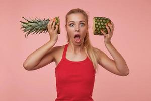 foto de estudio de una mujer hermosa y sorprendida con el pelo largo y rubio sosteniendo mitades de piña cerca de sus oídos, mirando la cámara con los ojos abiertos y la boca abierta