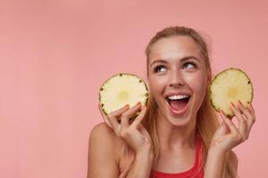 feliz joven mujer atractiva con maquillaje natural y peinado casual mirando alegremente hacia arriba, divirtiéndose con anillos de piña fresca, aislada sobre fondo rosa foto