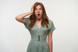 Indoor shot of surprised young woman with curly red hair, looking to camera with wide mouth opened, keeping palm on her head, isolated over white background photo