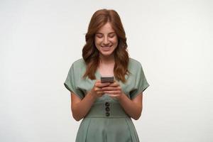 Indoor shot of pretty young lady in romantic dress holding smartphone in her hands, looking at screen and smiling cheerfully, isolated over white background photo