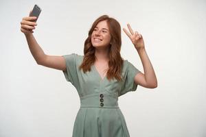 Studio shot of charming redhead young female making selfie with her smartphone, smiling to camera cheerfully and raising two fingers with peace gesture photo