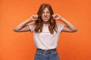 Studio photo of beautiful curly female in casual clothes grimacing and puckering face, closing ears with forefingers, trying to avoid loud sounds
