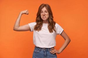 Studio shot of pretty redhead curly young woman showing her power in raised hand, wearing casual clothes, posing over orange background photo