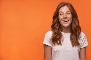 Young happy red haired female in white t-shirt looking aside with wide smile, smiling cheerfully and looking aside, isolated over orange background