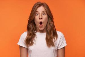 Close-up portrait of shocked curly female in white t-shirt posing over orange background with open-eyed face, raising eyebrows and opening mouth widely photo