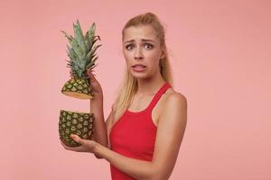 Studio shot of confused young pretty woman with casual hairstyle holding two pieces of pineapple, moving eyebrows and wrinkling forehead, isolated over pink background photo