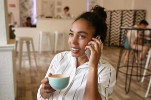 Beautiful young woman with bun haistyle posing over cafe interior with phone in hand, looking aside dreamily and smiling sincerely, holding cup of coffee