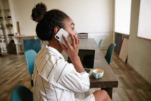 Indoor shot of attractive dark skinned woman with bun hairstyle sitting at long wooden table, talking on phone and looking thoughtfully out window photo