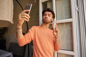 Indoor photo of handsome bearded man in peach color sweater making selfie with his smartphone