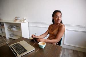 Charming dark skinned curly woman working at wooden table with modern laptop, writing notes in her daily planner, looking aside with light smile photo